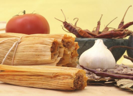 Tamales garnis avec une tomate, de l'ail et des piments secs sur un fond jaune.